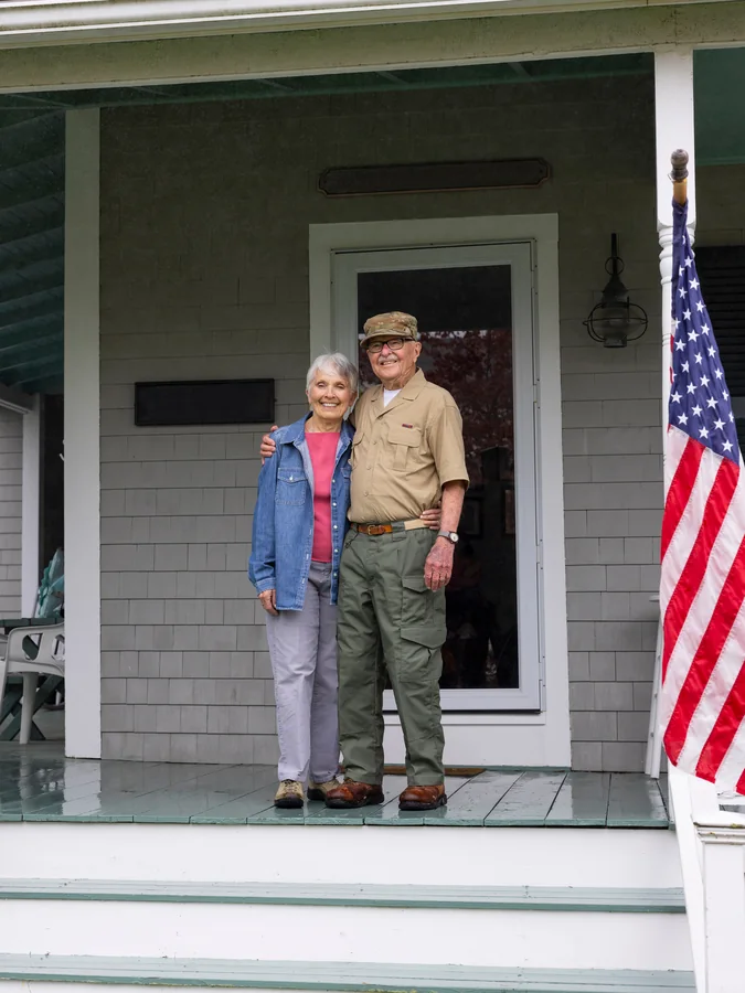 An older couple embracing on the porch of their home, an American flag displayed beside them